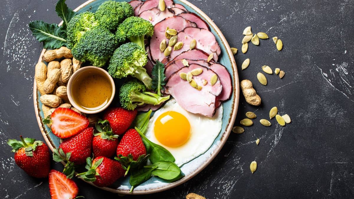 A balanced meal on a plate featuring slices of ham, a fried egg, broccoli, strawberries, spinach, and nuts with a small bowl of dressing.