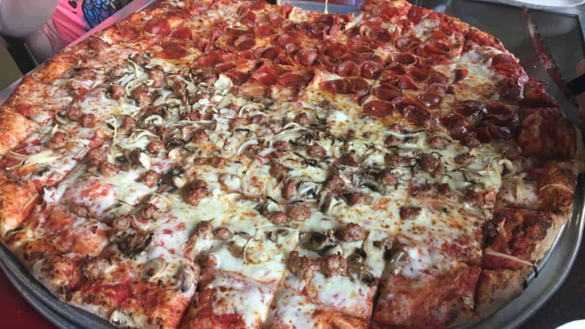 A large pizza, featured in extreme food eating contests, with half topped with pepperoni and the other half with mushrooms and cheese, displayed on a metal tray at a table.