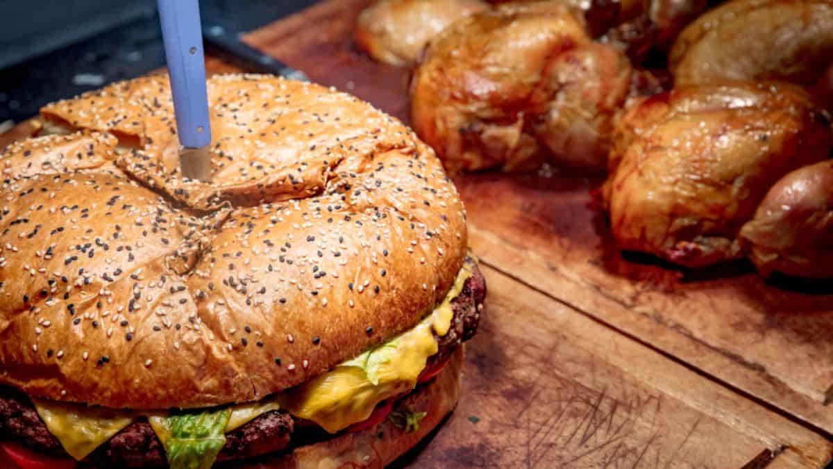 Large burger with sesame seed bun and knife inserted on top, on a wooden cutting board; roasted chickens in the background.