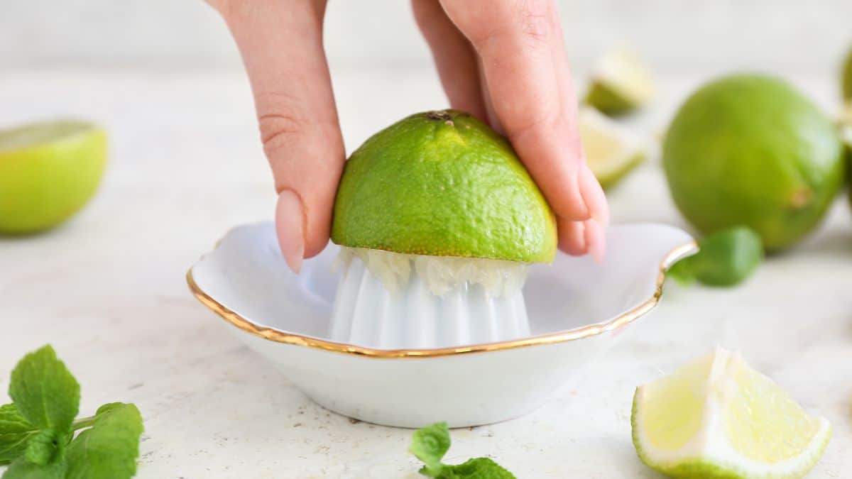 A person squeezing a lime on a citrus juicer with fresh limes and mint leaves on the countertop.