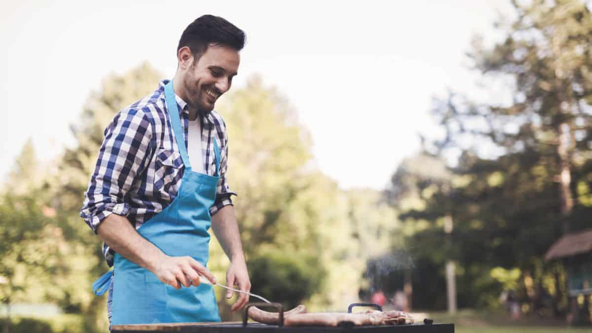 A man in a blue apron and plaid shirt smiles while grilling outdoors in a sunny park setting.