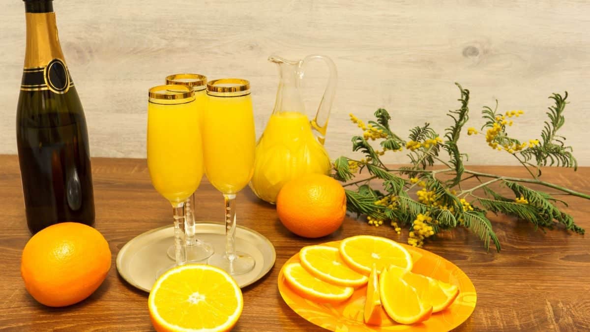 A still life composition featuring mimosas in flute glasses, a bottle of champagne, fresh oranges, and a pitcher on a wooden table with greenery in the background.