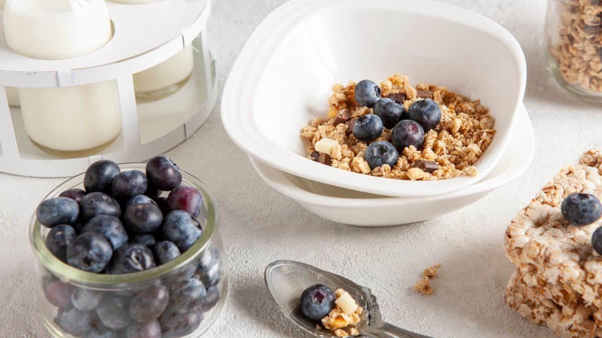 A bowl of granola topped with blueberries, surrounded by more blueberries, granola bars, and yogurt containers on a light textured surface.