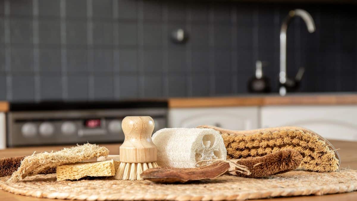 An array of eco-friendly kitchen cleaning tools, including wooden brushes and natural sponges, displayed on a woven mat with a modern kitchen backdrop.