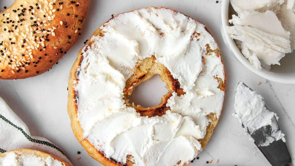 A bagel with cream cheese, with a bite taken out of it, alongside more cream cheese and a spreading knife.