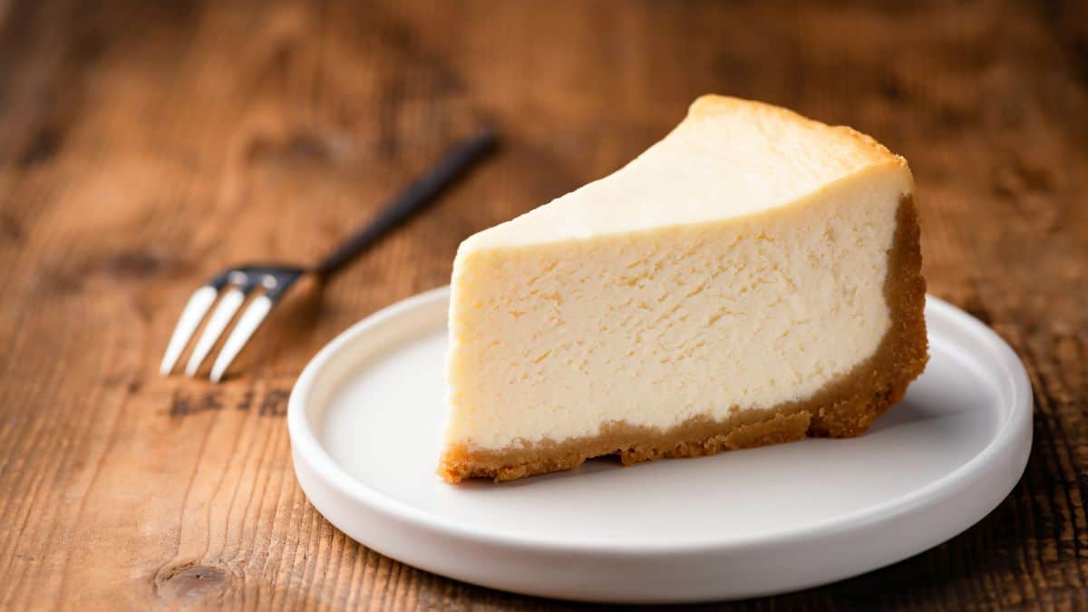 A slice of classic cheesecake on a white plate with a fork on a wooden table.