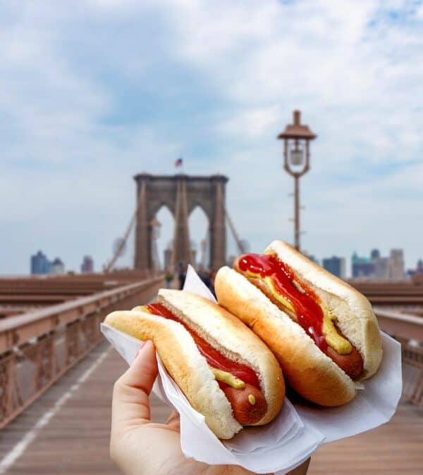 A hand holding two hot dogs with a view of brooklyn bridge in the background.