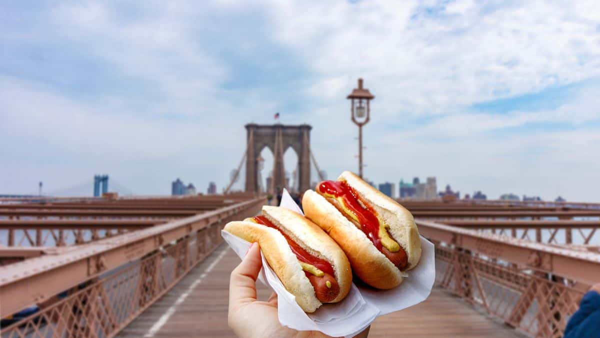 A hand holding two hot dogs with a view of brooklyn bridge in the background.