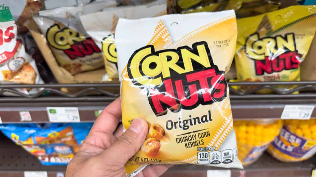 A hand holding a bag of corn nuts original flavor in a grocery store aisle.
