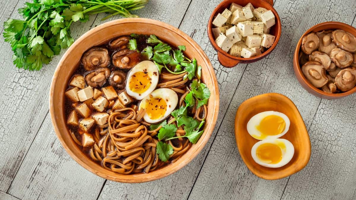 A bowl of ramen with tofu, mushrooms, soft-boiled eggs, and greens, accompanied by side dishes.