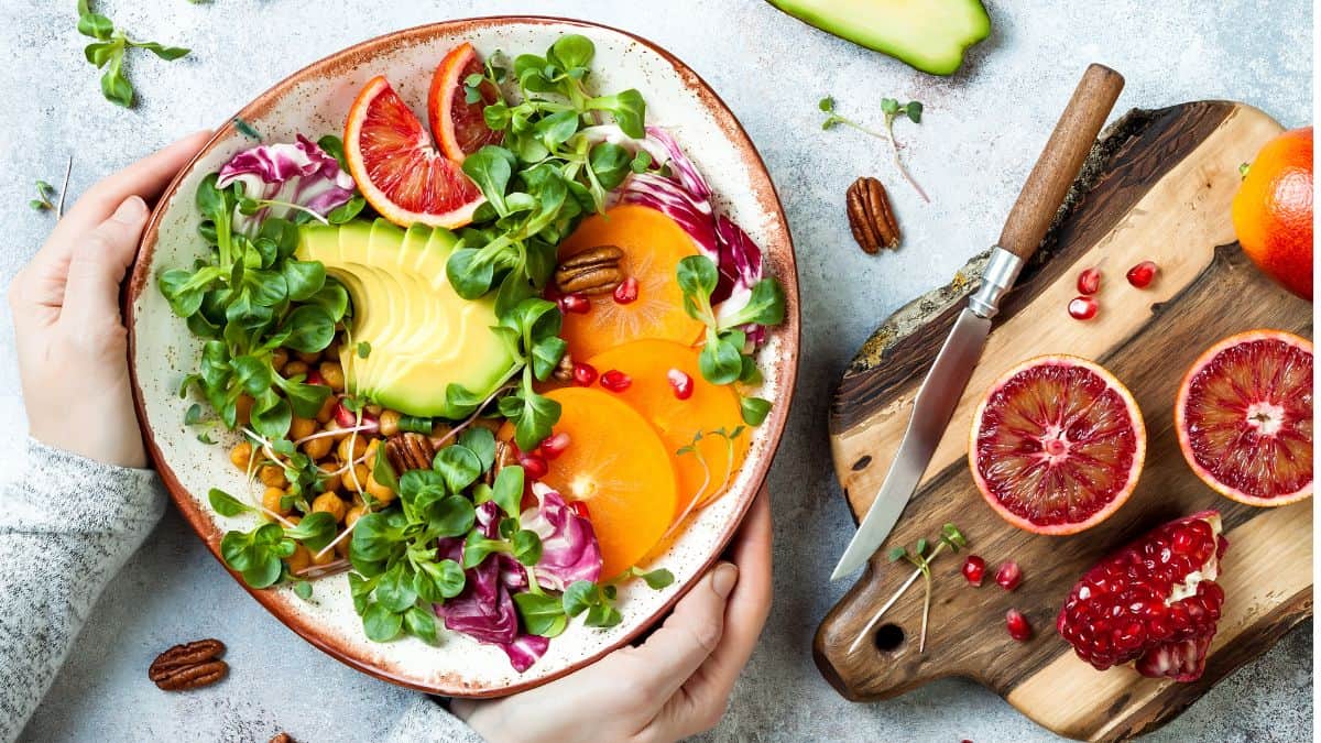 Hands holding a colorful salad bowl with avocado, blood oranges, and greens, next to a cutting board with fruit and nuts.