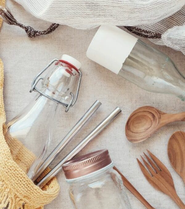 Assorted reusable items including glass bottles, wooden utensils, fabric bags, and metal containers arranged on a light surface.