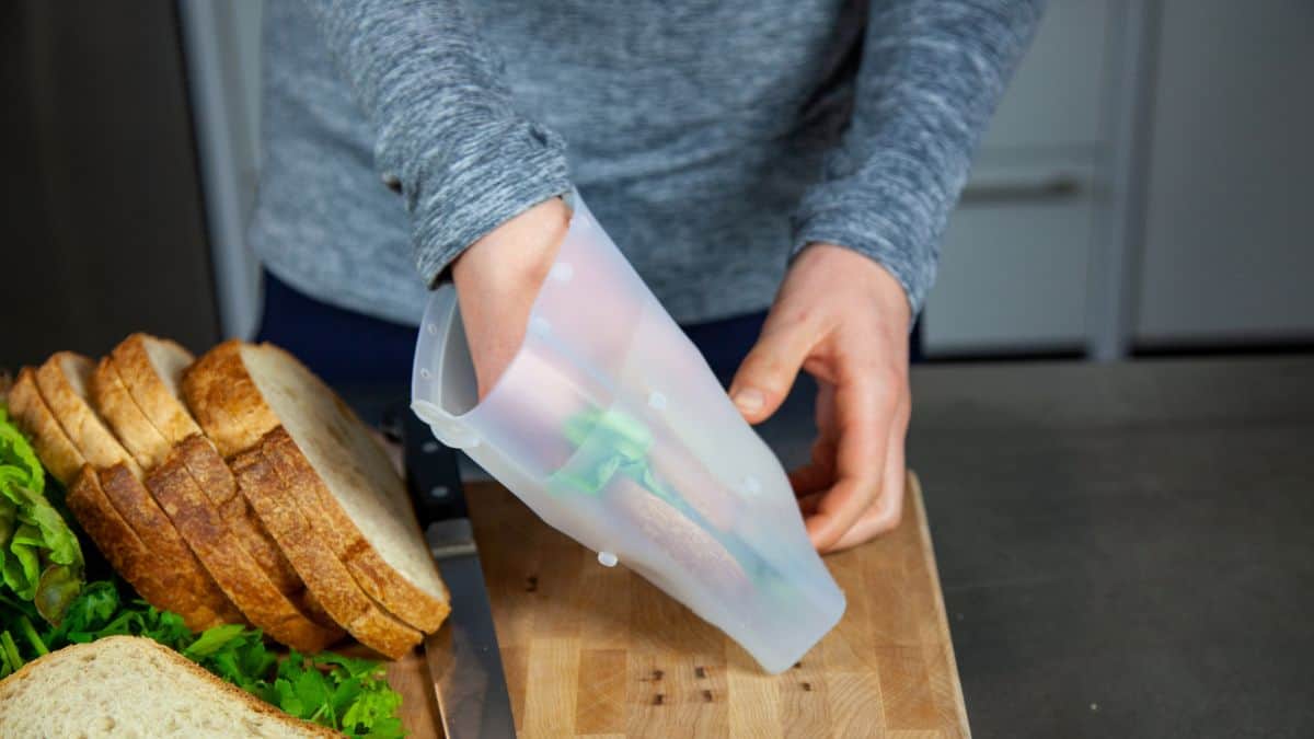 A person pours mixed vegetables from a plastic container onto a wooden cutting board next to sliced bread.