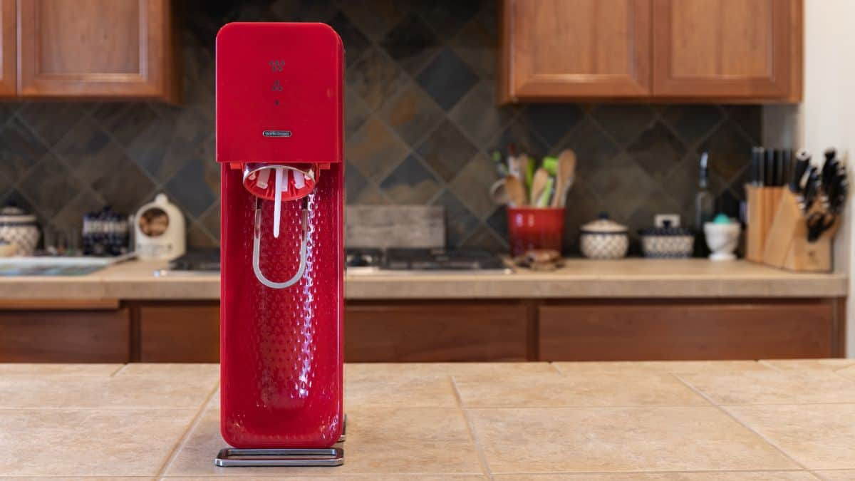 A red soda maker machine on a kitchen counter with wooden cabinets in the background.