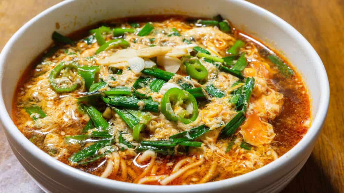 A bowl of spicy noodle soup garnished with green onions, sliced peppers, and egg, served on a wooden table.