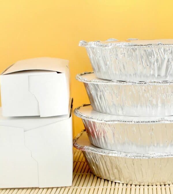 Stacks of aluminum takeout containers and white cardboard boxes on a bamboo mat against a yellow background.