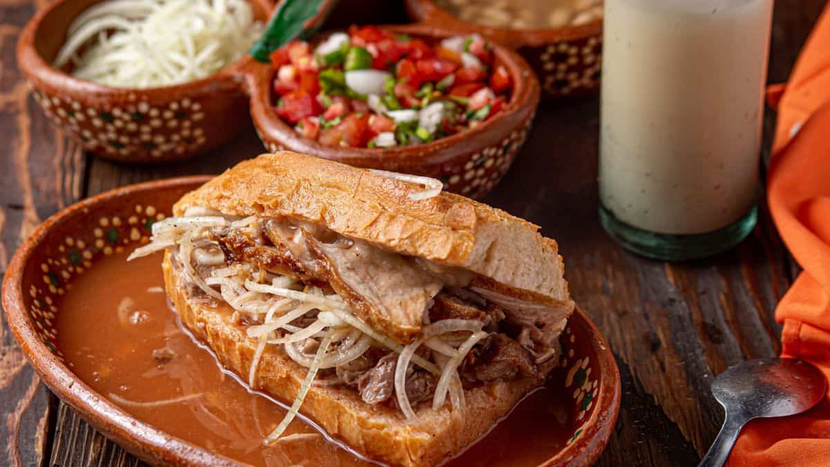 Traditional mexican torta ahogada with a side of salsa and a creamy beverage.