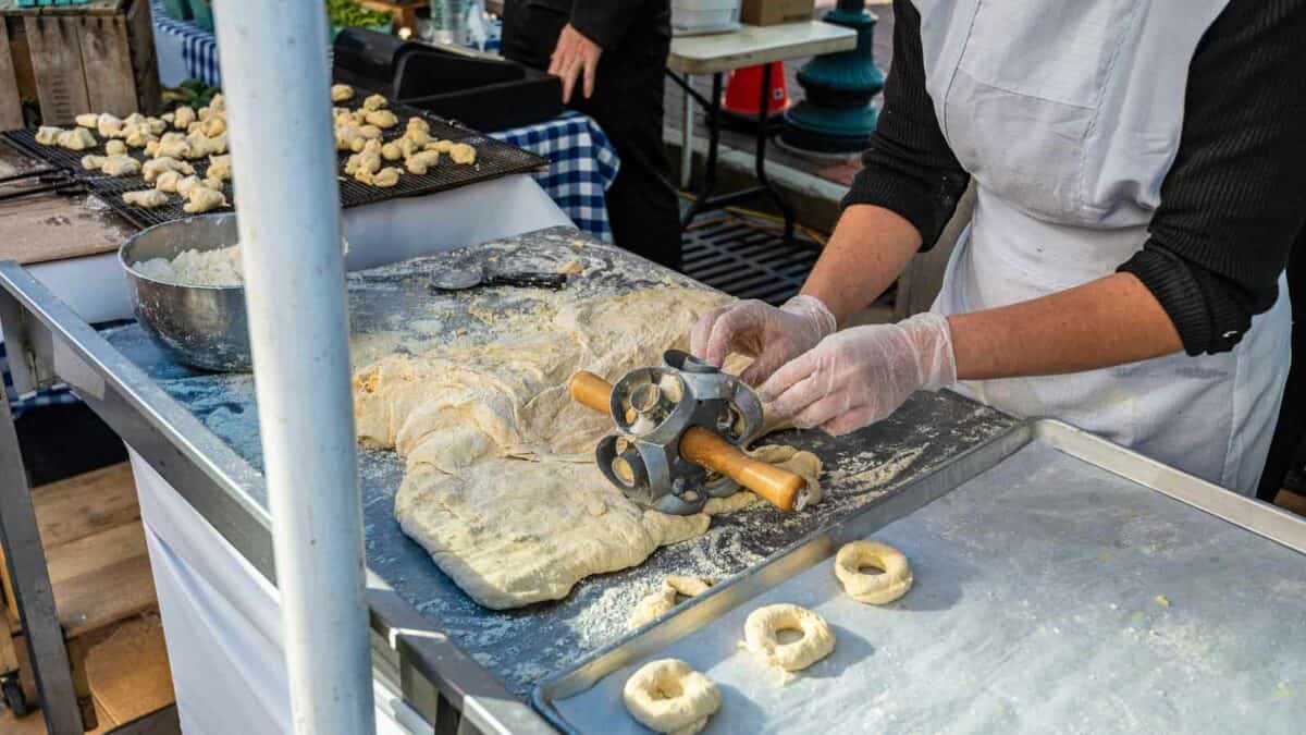 A person in an apron using a dough cutter to make bagels on a floured table at a street market stall.