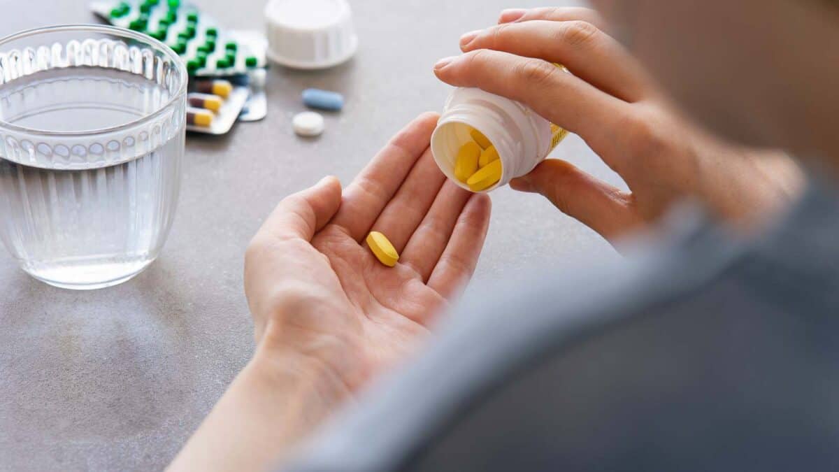 A person is pouring pills from a bottle into their hand with a glass of water and more medication in the background.