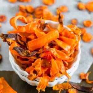 A bowl of homemade carrot chips on a gray surface, showcasing crispy, orange slices with slightly charred edges.