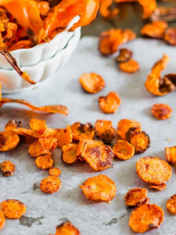 A bowl of crispy carrot chips next to scattered chips on parchment paper.