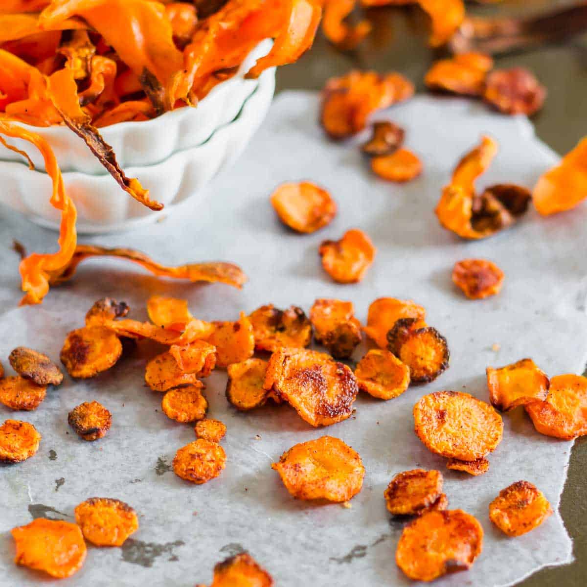 A bowl of crispy carrot chips next to scattered chips on parchment paper.