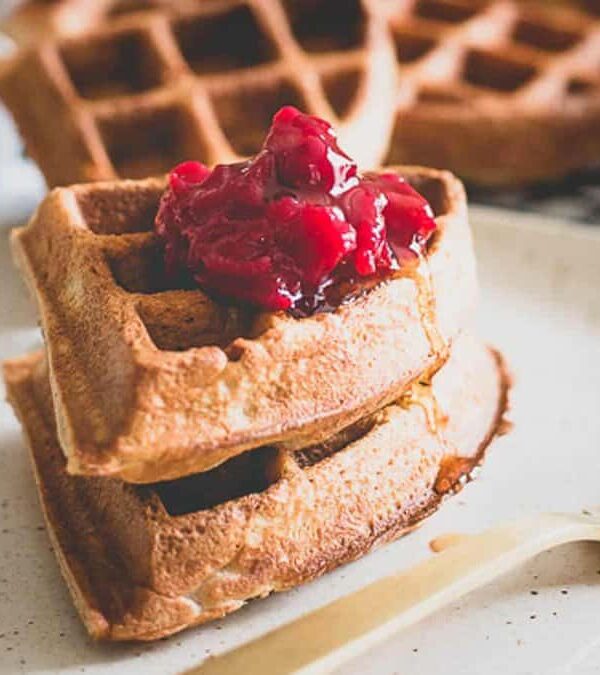 Stack of whole grain waffles topped with raspberry compote on a white plate, with a fork on the side.
