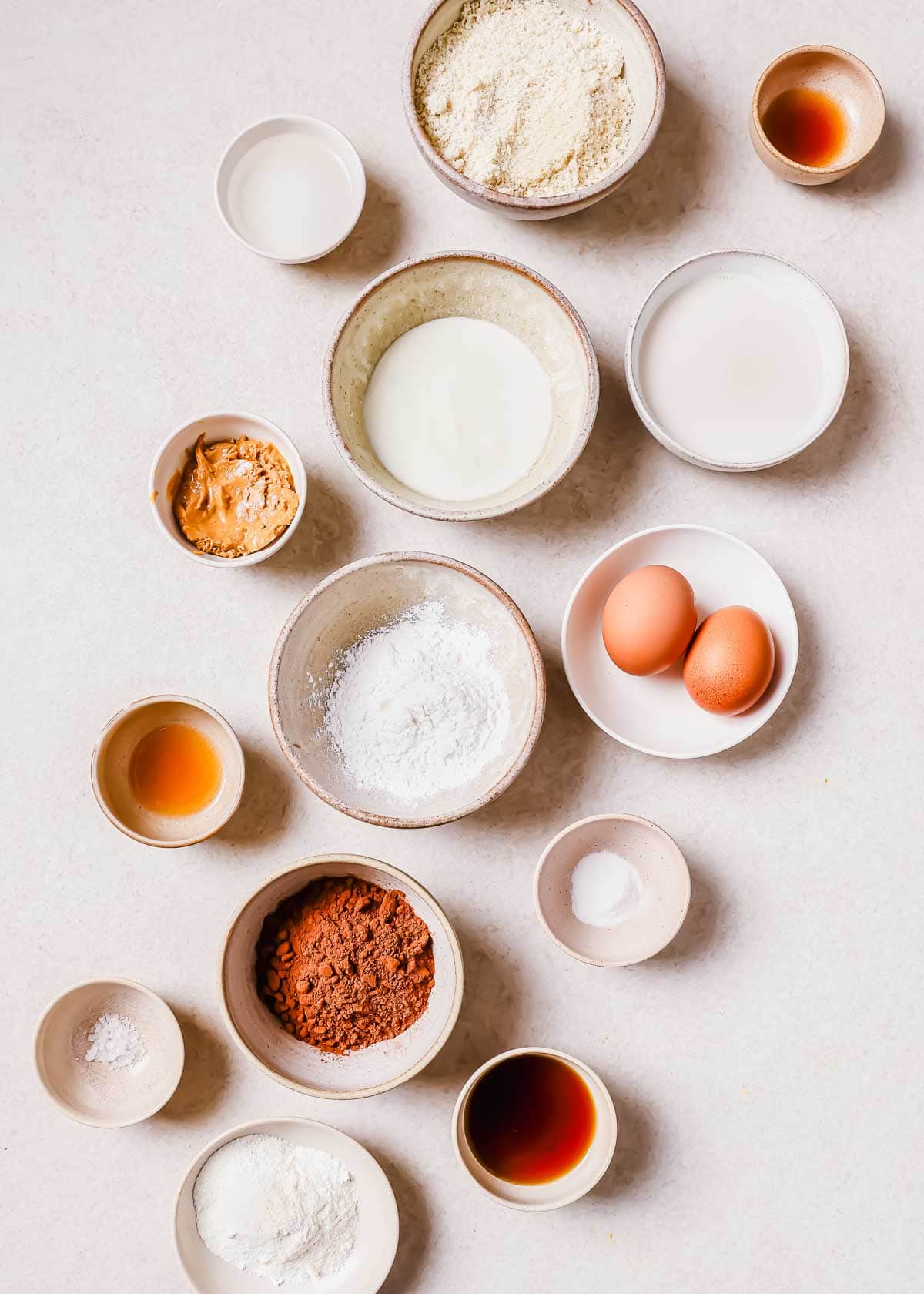 Various baking ingredients neatly arranged in bowls on a light surface, including flour, eggs, milk, cocoa, and sugar.