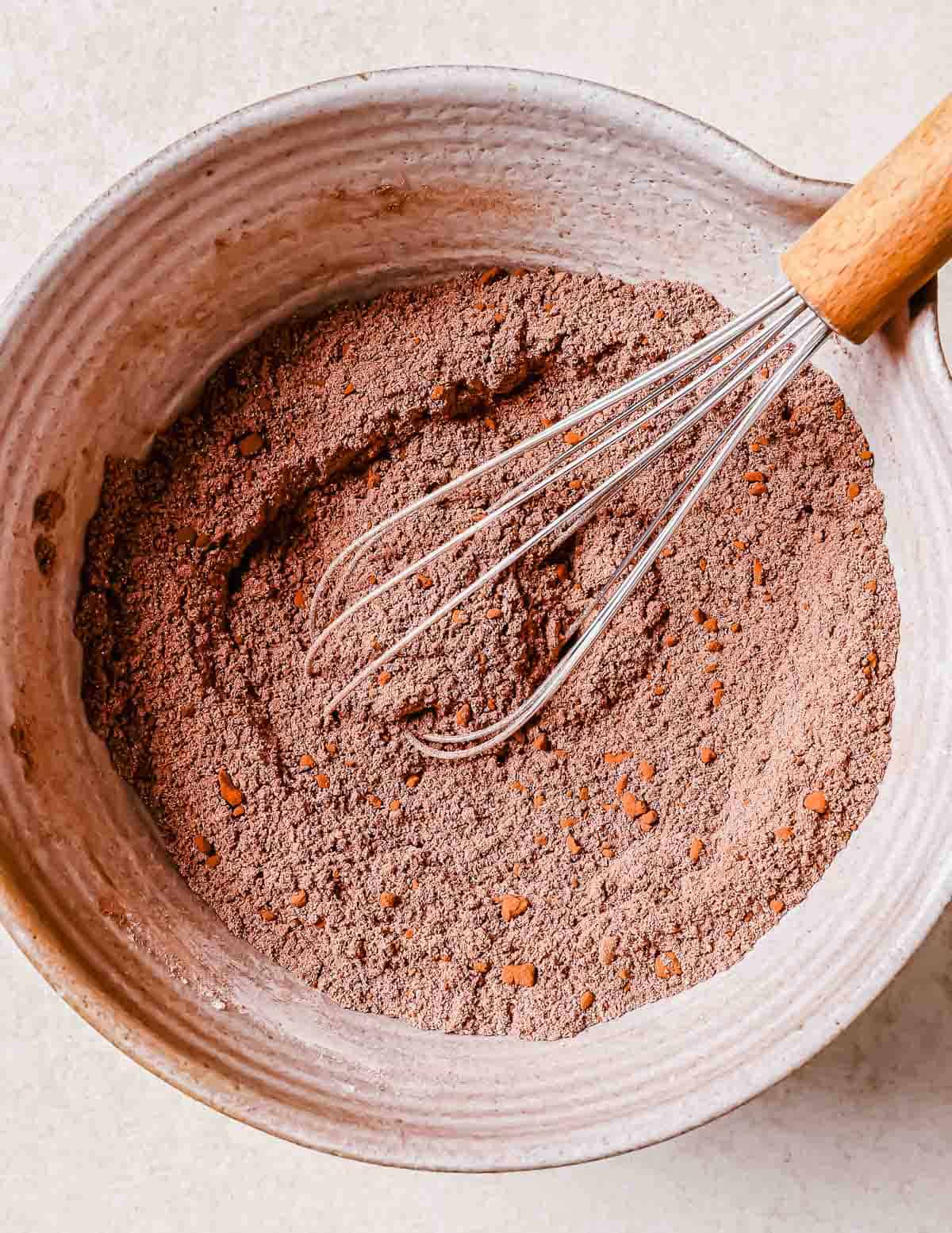 A bowl of dry cocoa powder mixed with flours, with a metal whisk inside, placed on a light beige surface.