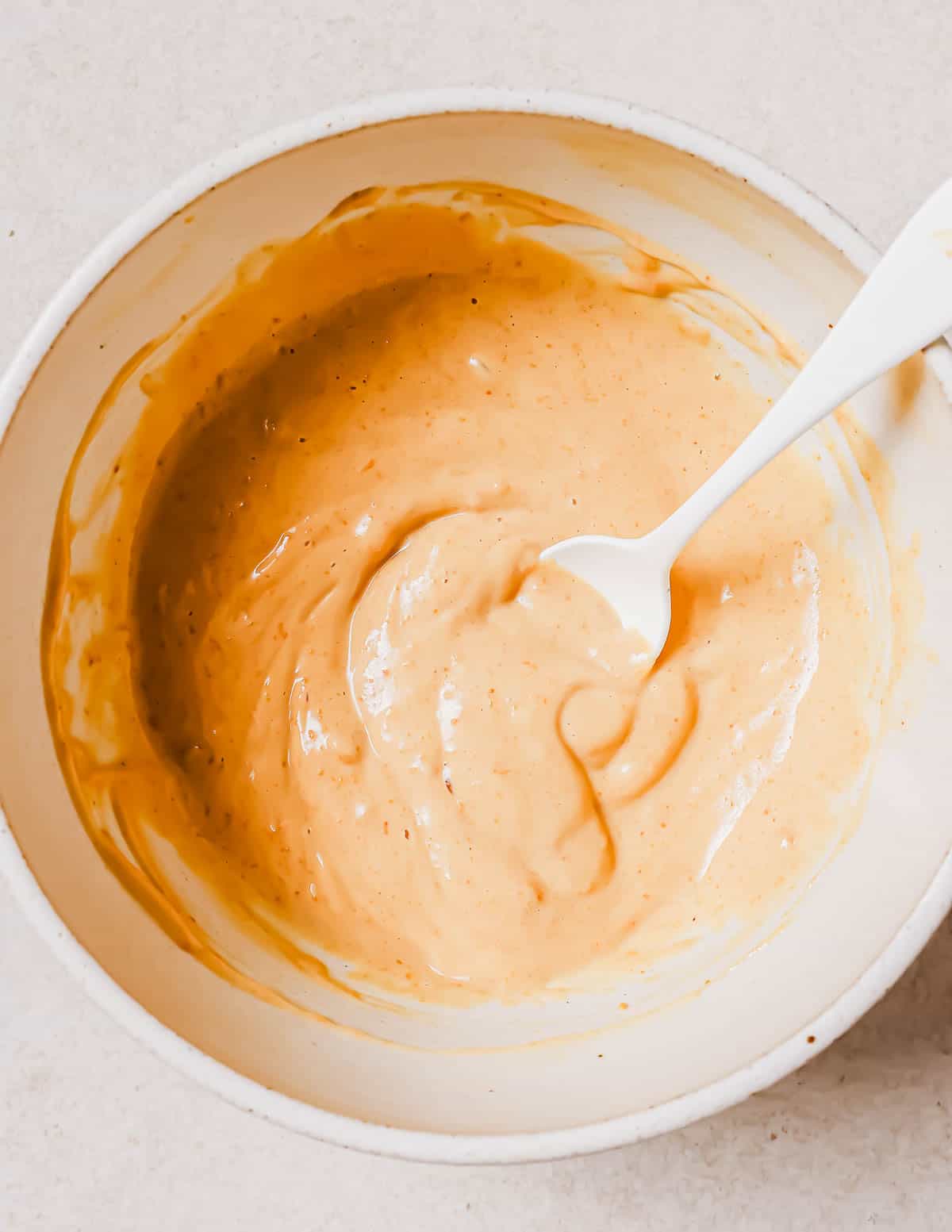 A bowl of creamy waffle batter with a white spoon, on a light beige surface.