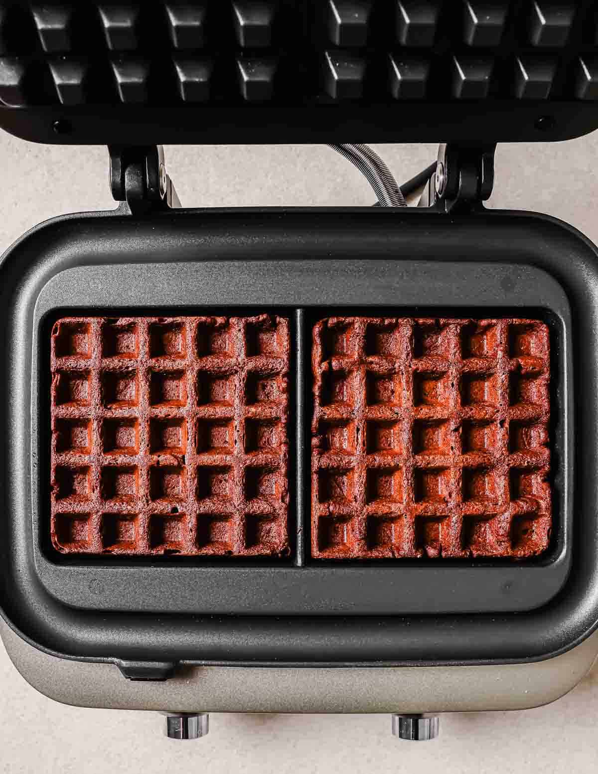 Two freshly cooked chocolate waffles in an open waffle iron, showing golden-brown surfaces with deep indentations.