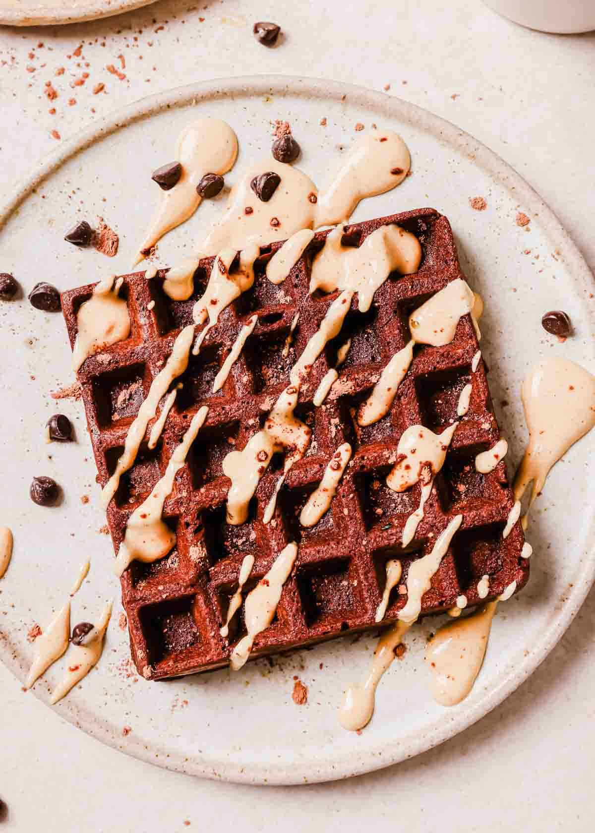 A chocolate waffle topped with peanut butter sauce and chocolate chips on a speckled plate.