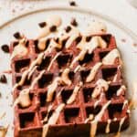 A square chocolate waffle drizzled with peanut butter glaze and sprinkled with chocolate chips on a white plate.