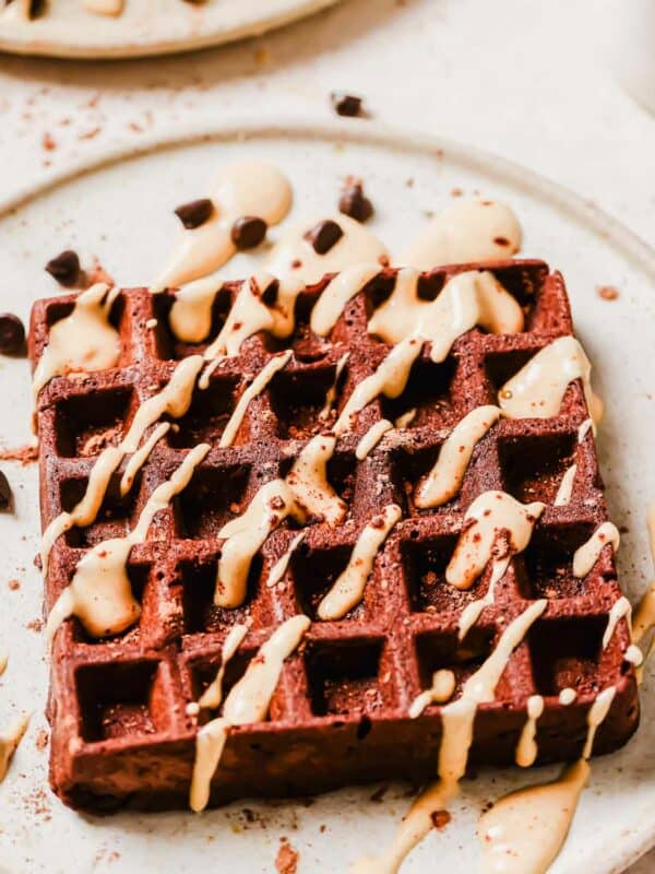 A square chocolate waffle drizzled with peanut butter glaze and sprinkled with chocolate chips on a white plate.