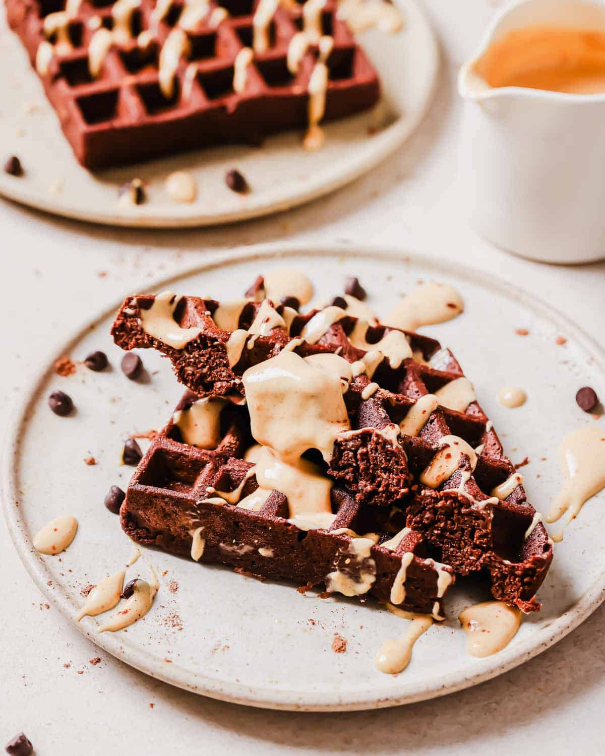 Chocolate waffles topped with peanut butter sauce on a white plate, garnished with chocolate chips, served with a side of more sauce.