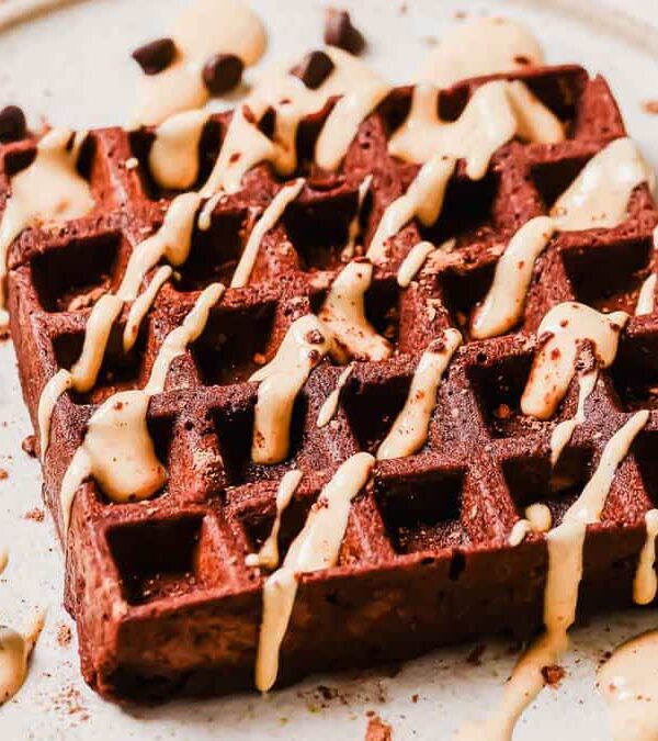 A chocolate waffle topped with peanut butter drizzle and chocolate chips on a ceramic plate.