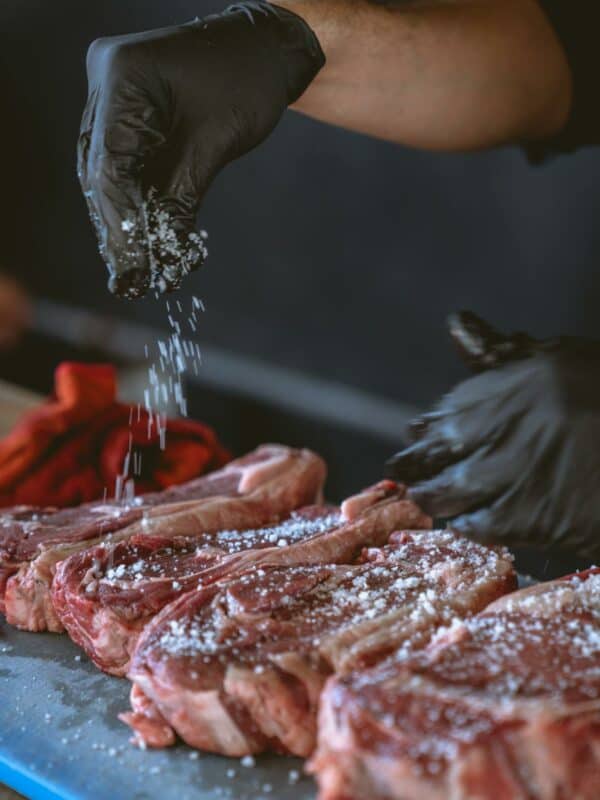 A person wearing black gloves seasoning a chuck eye steak with salt.