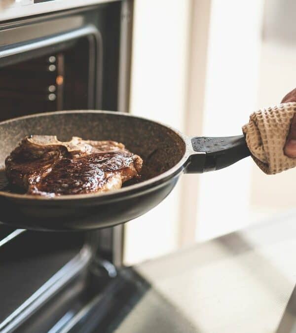A person holds a frying pan with steak, taking it out of an oven.