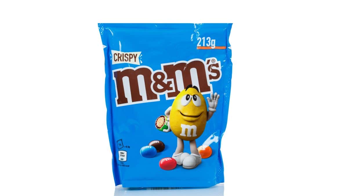 A bag of crispy m&m's with the yellow m&m character peeking out, holding a candy, isolated on a white background.