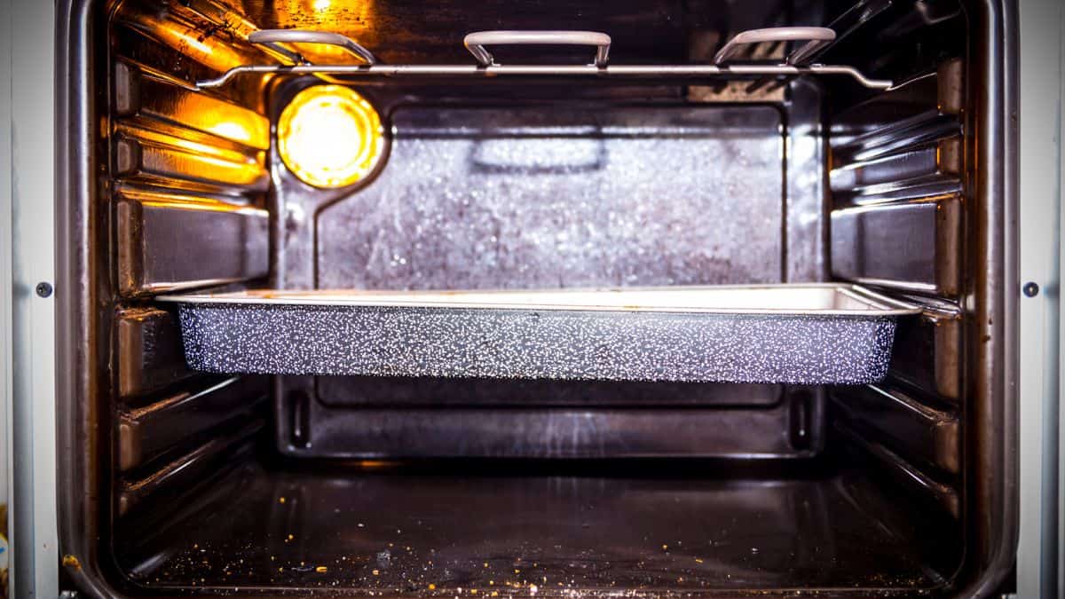 Interior of an oven with a light on and an empty baking tray inside, showing metal racks and reflective surfaces.