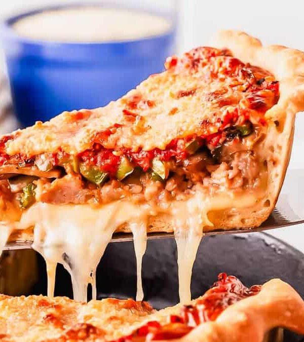 A slice of deep-dish pizza being lifted, showing melty cheese and vegetable toppings, with more pizza and a salad in the background.