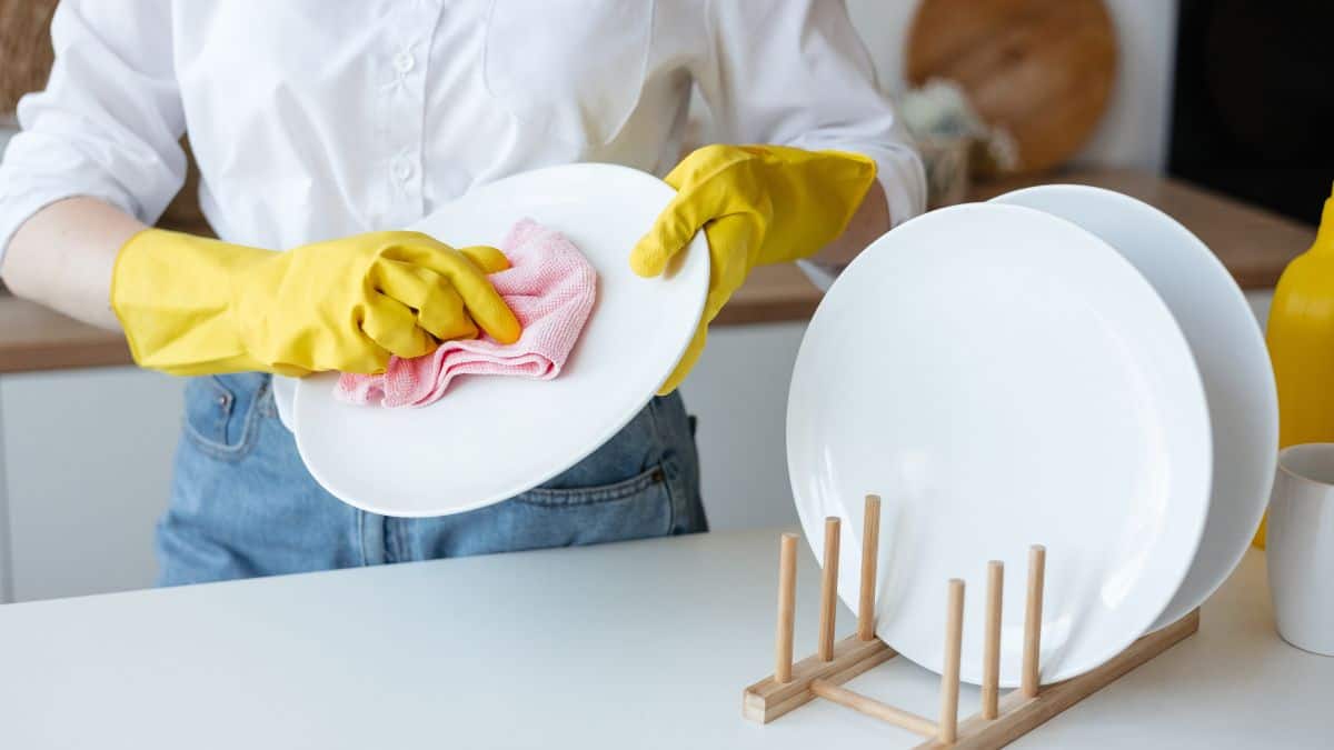 Person in yellow gloves wiping a white plate with a pink cloth at a kitchen table, with more plates and cleaning supplies nearby.