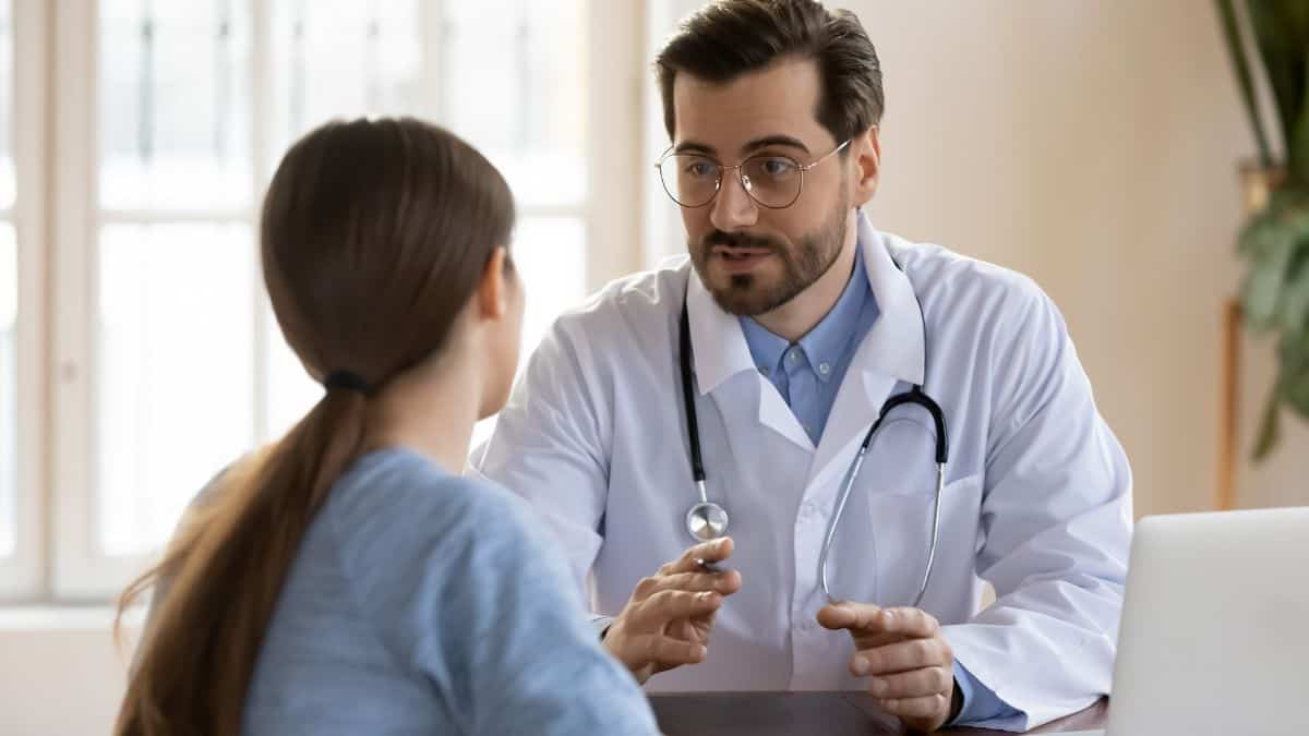 A male doctor in glasses and a stethoscope engaged in conversation with a female patient in an office setting.
