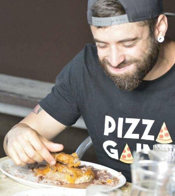 Man in a "pizza gang" t-shirt smiles while eating ribs at a table, with a smartphone showing a timer reading 9 minutes, 11 seconds.