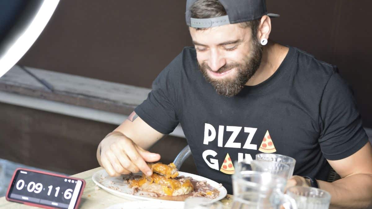 Man in a "pizza gang" t-shirt smiles while eating ribs at a table, with a smartphone showing a timer reading 9 minutes, 11 seconds.