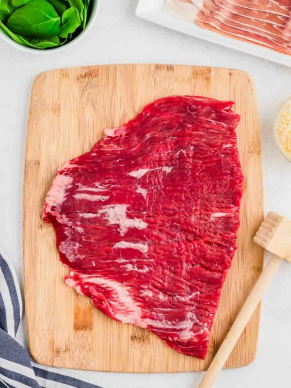 Raw flank steak on a wooden cutting board with cooking ingredients in the background.