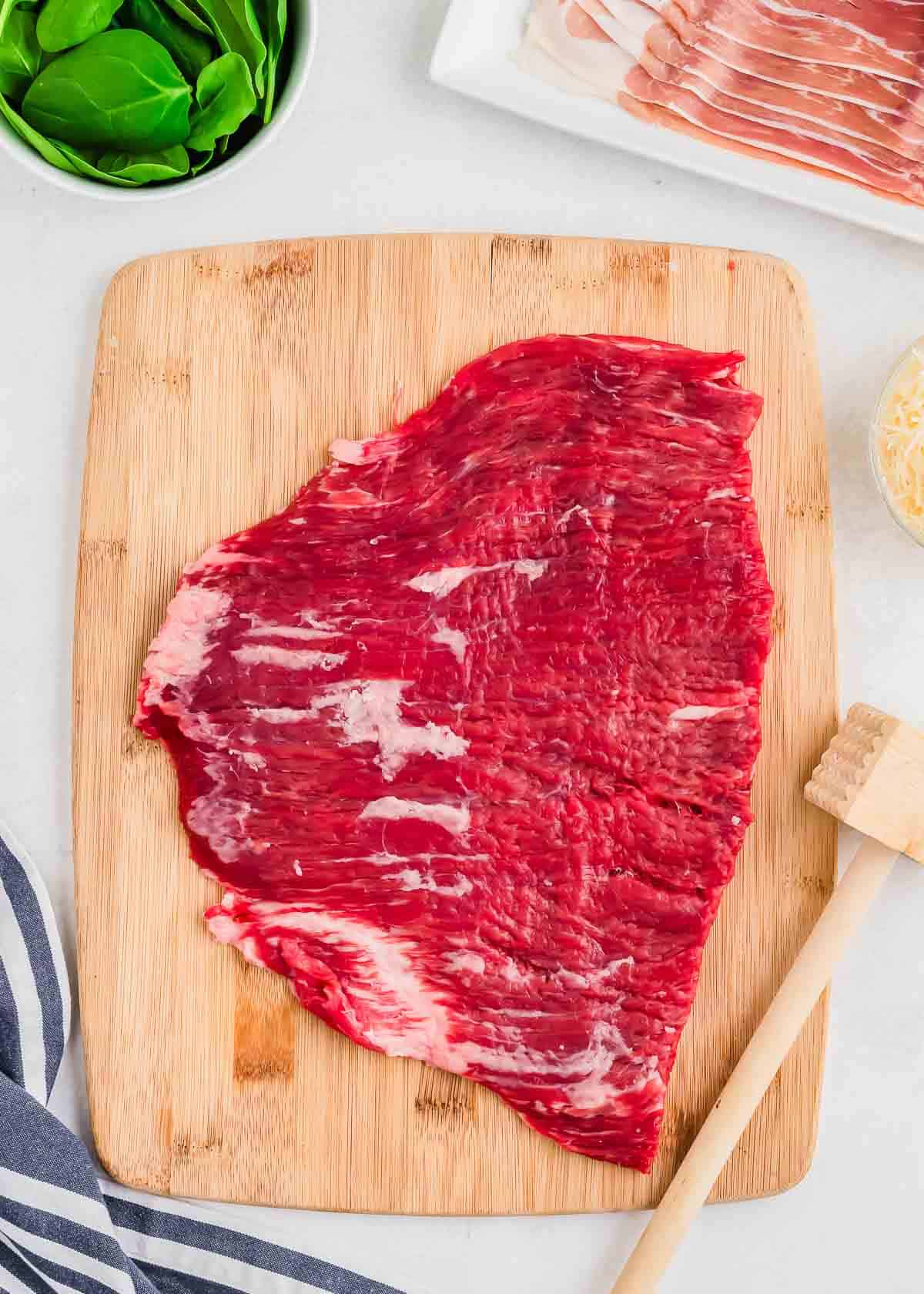 Raw flank steak on a wooden cutting board with cooking ingredients in the background.