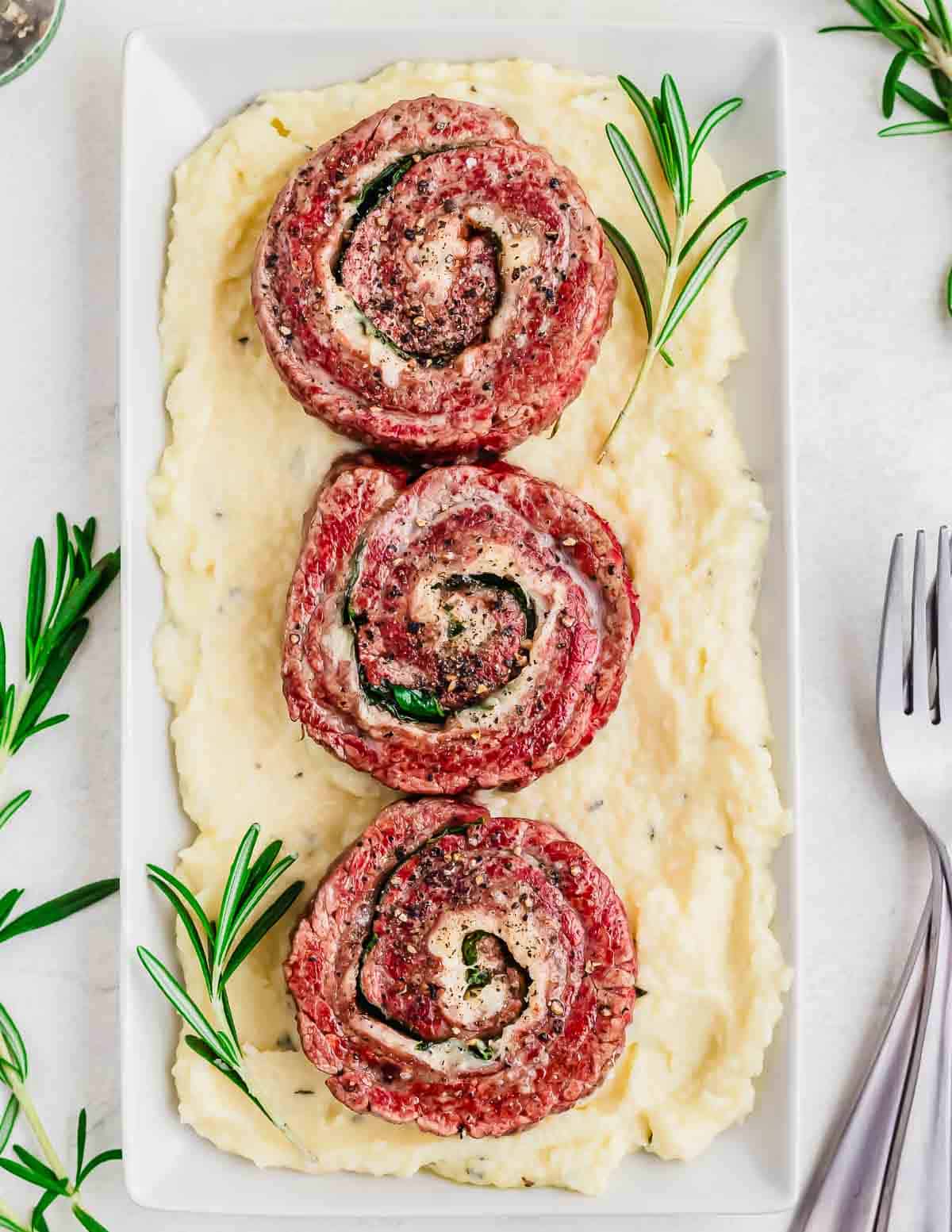 Beef flank steak pinwheels on a bed of mashed parsnips garnished with rosemary sprigs.
