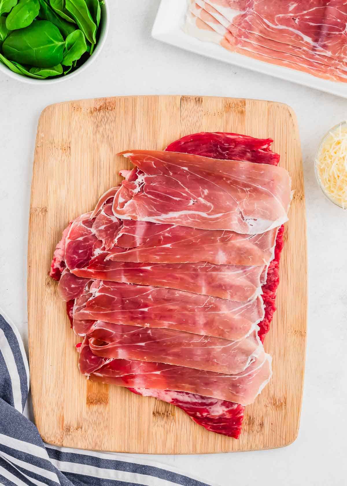 Thin slices of prosciutto arranged on top of pounded flank steak on a wooden cutting board, with a bowl of greens and grated cheese nearby.