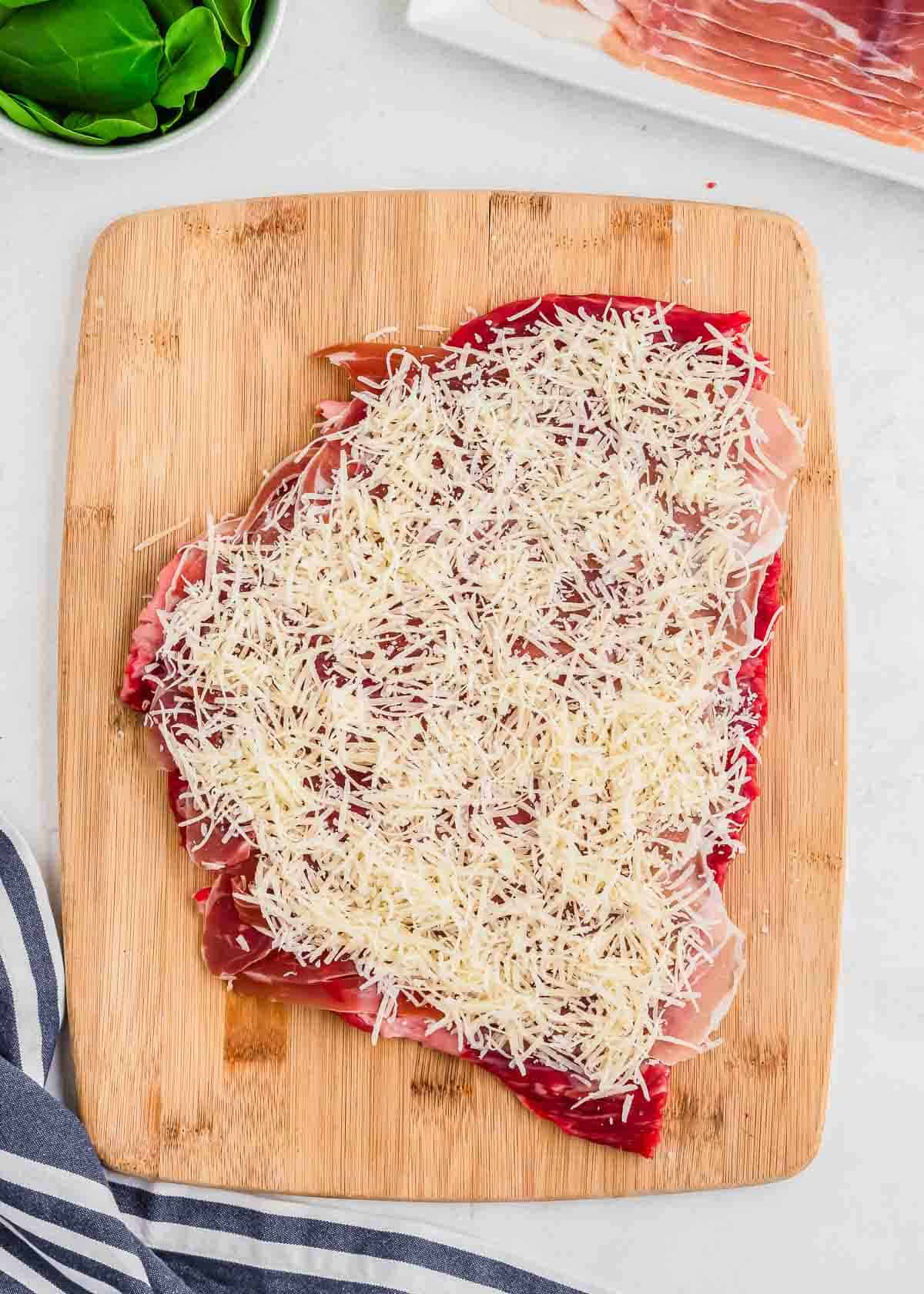 A cutting board with flank steak layered with shredded cheese, ready for cooking.